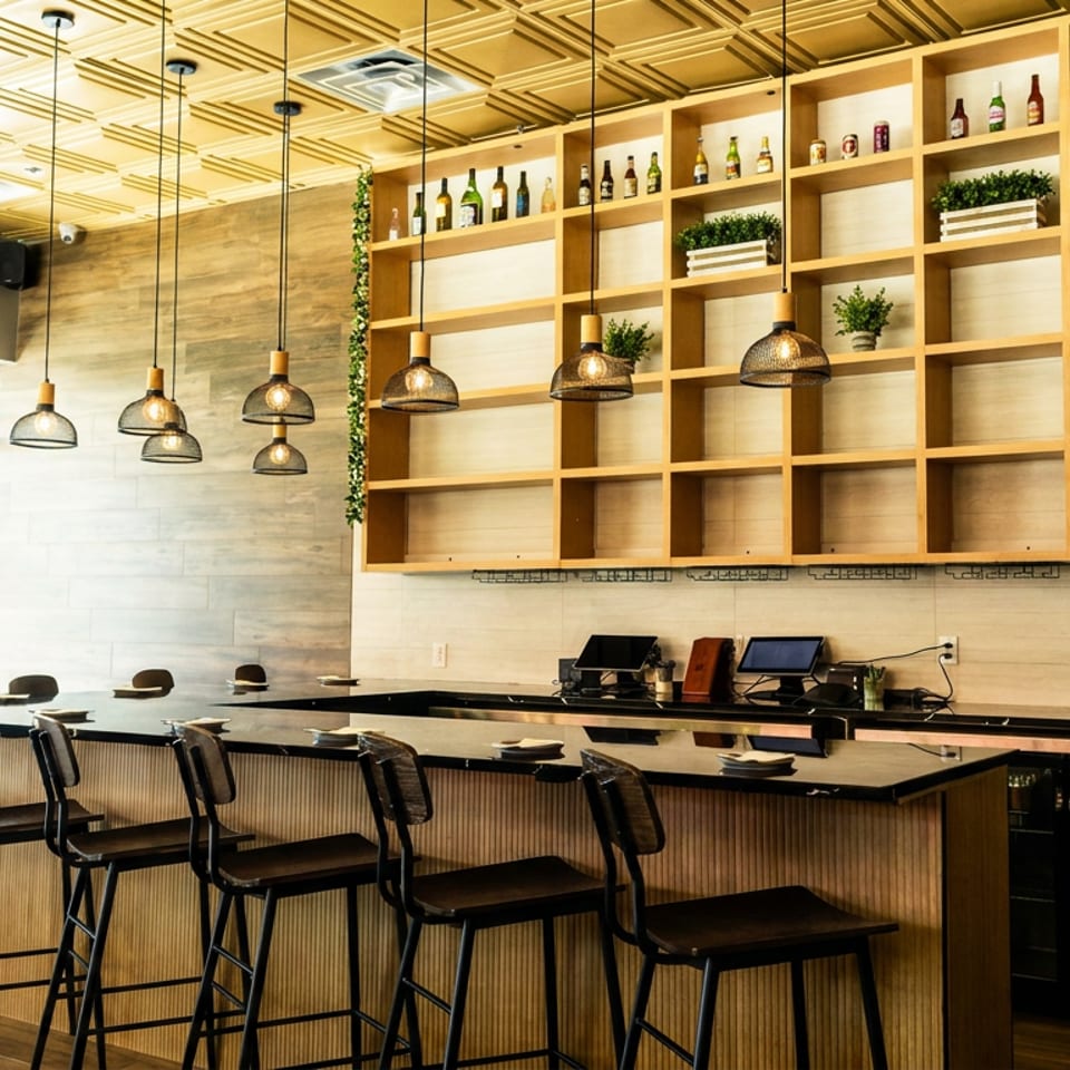 Bright modern restaurant interior with a bar counter, pendant lights, shelves displaying bottles and plants, and window seating.