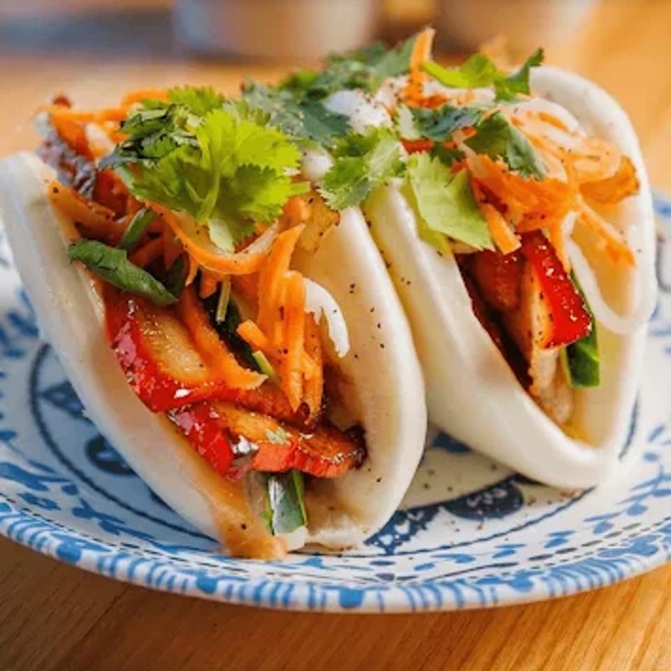 Two soft tacos filled with grilled meat, red peppers, carrots, herbs, and cilantro on a blue-and-white patterned plate.