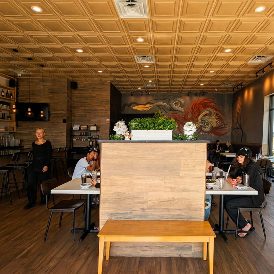Warmly lit modern restaurant interior with a bar, seated diners, wooden furnishings, large windows, and colorful wall mural.